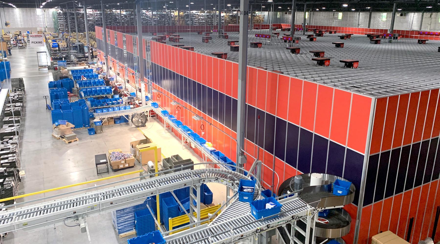 Interior of a large warehouse featuring an automated storage and retrieval system with numerous red and black storage bins arranged in a grid. Blue bins are visible on conveyor belts and workstations along the perimeter. The warehouse is well-lit with high ceilings, and various sections are labeled for organization.